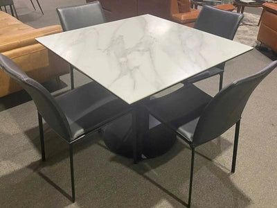 Square ceramic top Prisma Dining Table with matte black base and four chairs in modern setting.