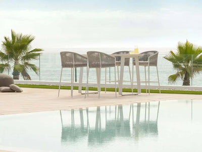 Outdoor dining area featuring Jiselle Outdoor Bar Chairs with white frames and grey rope, overlooking the ocean.