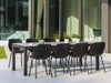Dante Outdoor Ceramic Dining Table in modern patio setting with black chairs and greenery.