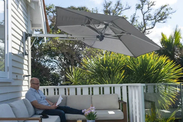 Retreat Wall-Mounted Outdoor Umbrella in grey providing shade over a man sitting on a beige outdoor sofa on a balcony